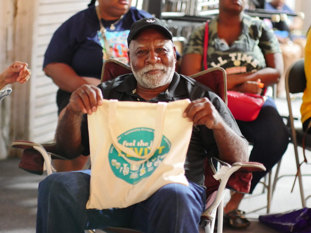 Older man with white beard and hat on holds up canvas bag during concert series.