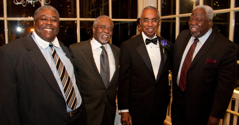 Four middle aged black men in suits and ties smile for a photo together at a formal event.